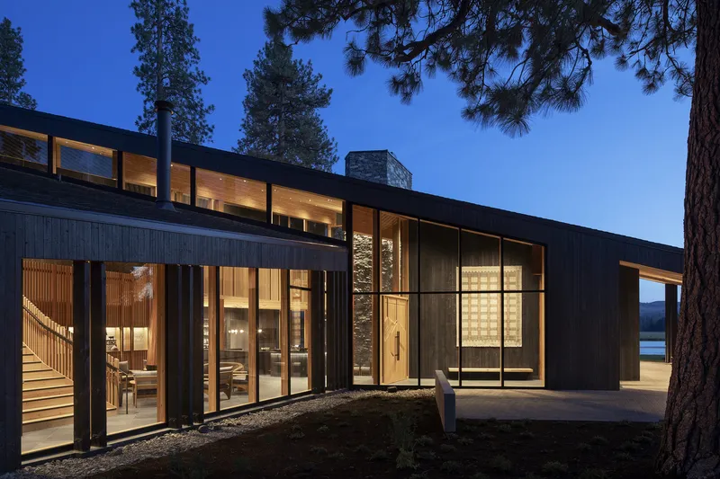Night view of the black timber-clad facade with illuminated interiors visible through expansive glass walls beneath pines
