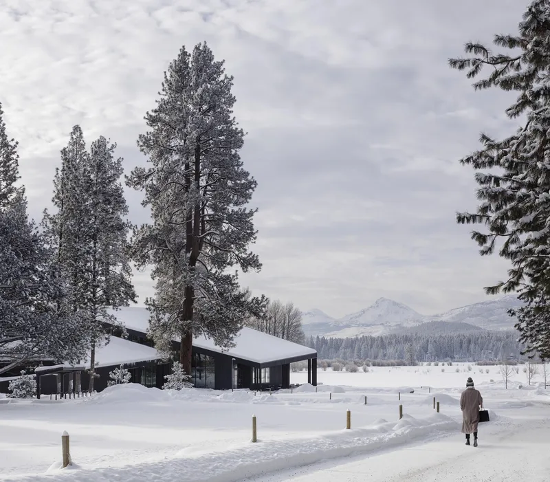 Low-slung black-clad volume in a snowy landscape with a figure walking along a cleared path toward mountains
