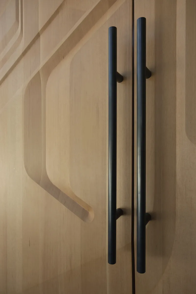 Close-up of black cylindrical pulls on light timber cabinetry with carved panel detail in afternoon shadow