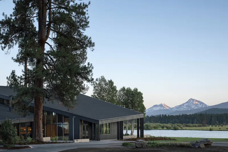 Low pavilion with dark metal cladding beside a lake with snow-capped peaks at sunset