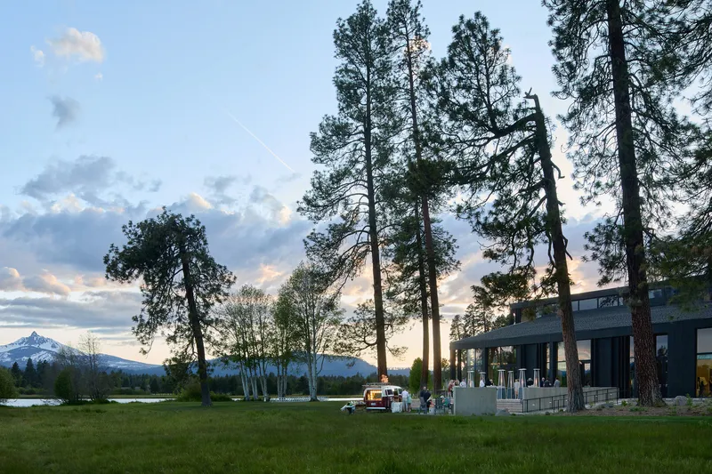 Exterior terrace with scattered visitors beneath towering pines at dusk, overlooking a lake and distant mountains