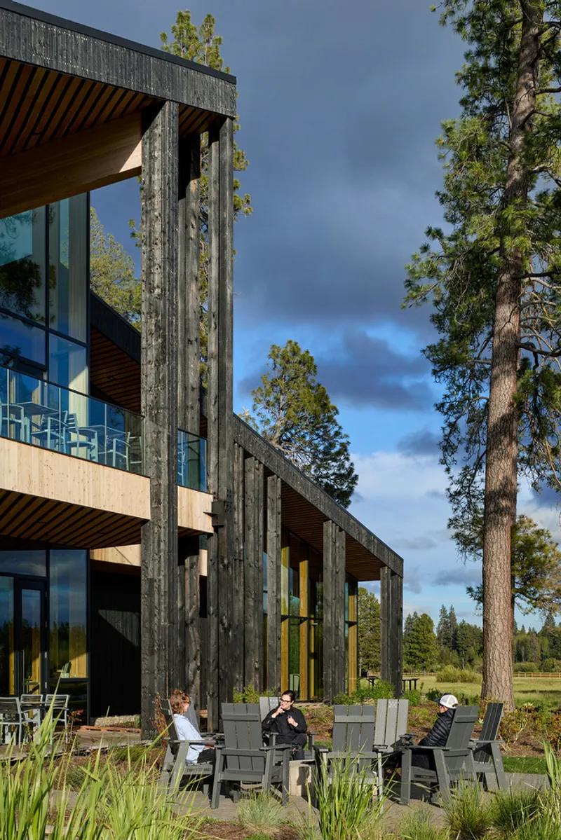 Charred timber facade with floor-to-ceiling glazing and cantilevered balcony, framed by tall pines and guests seated below