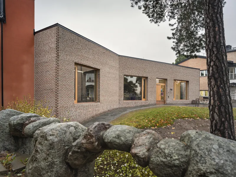 Curved brick volume with timber-framed openings beyond a stone retaining wall and pine tree