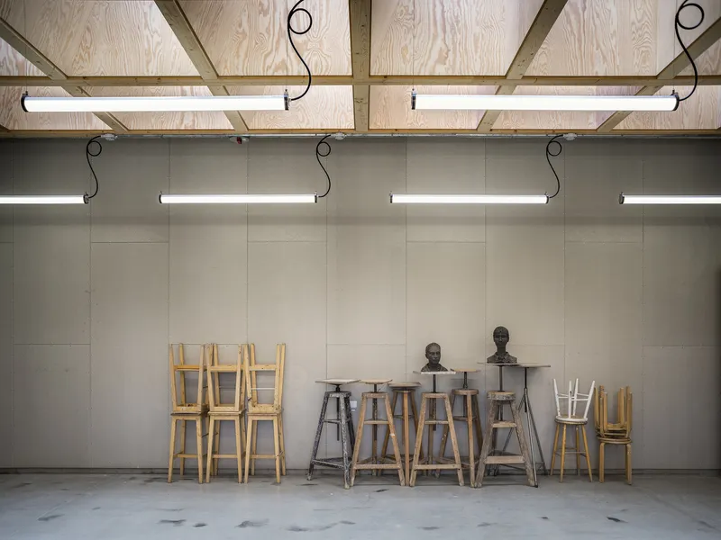 Studio wall with plywood ceiling beams, hanging fluorescent lights, and wooden stools arranged along concrete floor