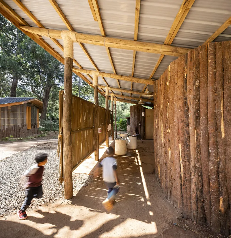 Covered walkway with exposed eucalyptus log columns and translucent roofing as children run through filtered sunlight