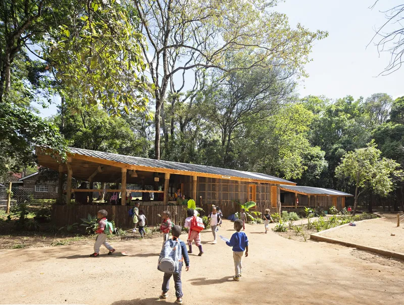 Long facade with corrugated panels and metal roof canopy as children with backpacks walk across the clearing