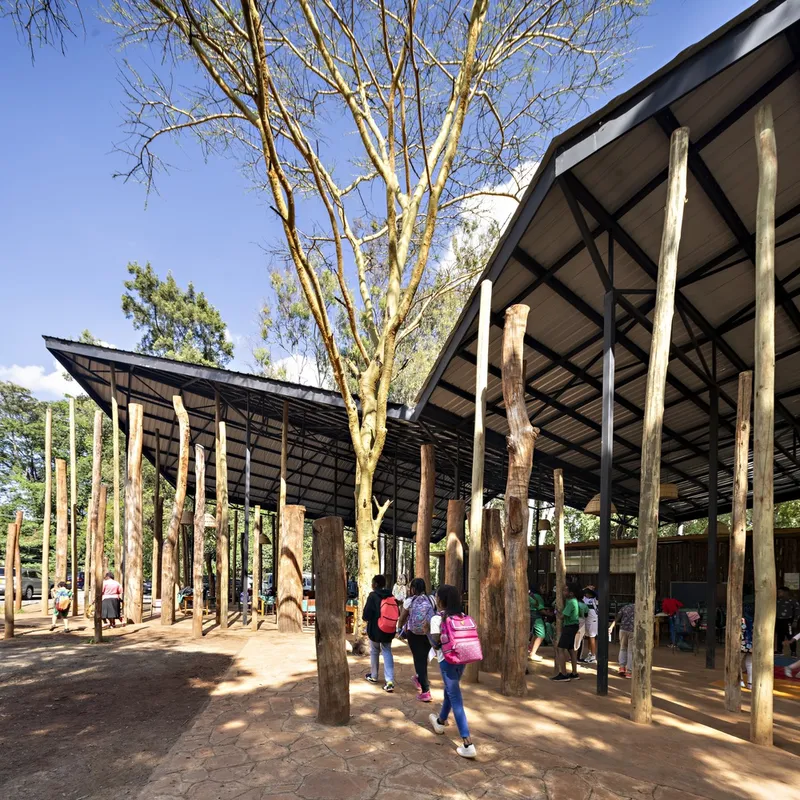 Covered courtyard with eucalyptus tree trunk columns and children with backpacks walking beneath a black steel roof