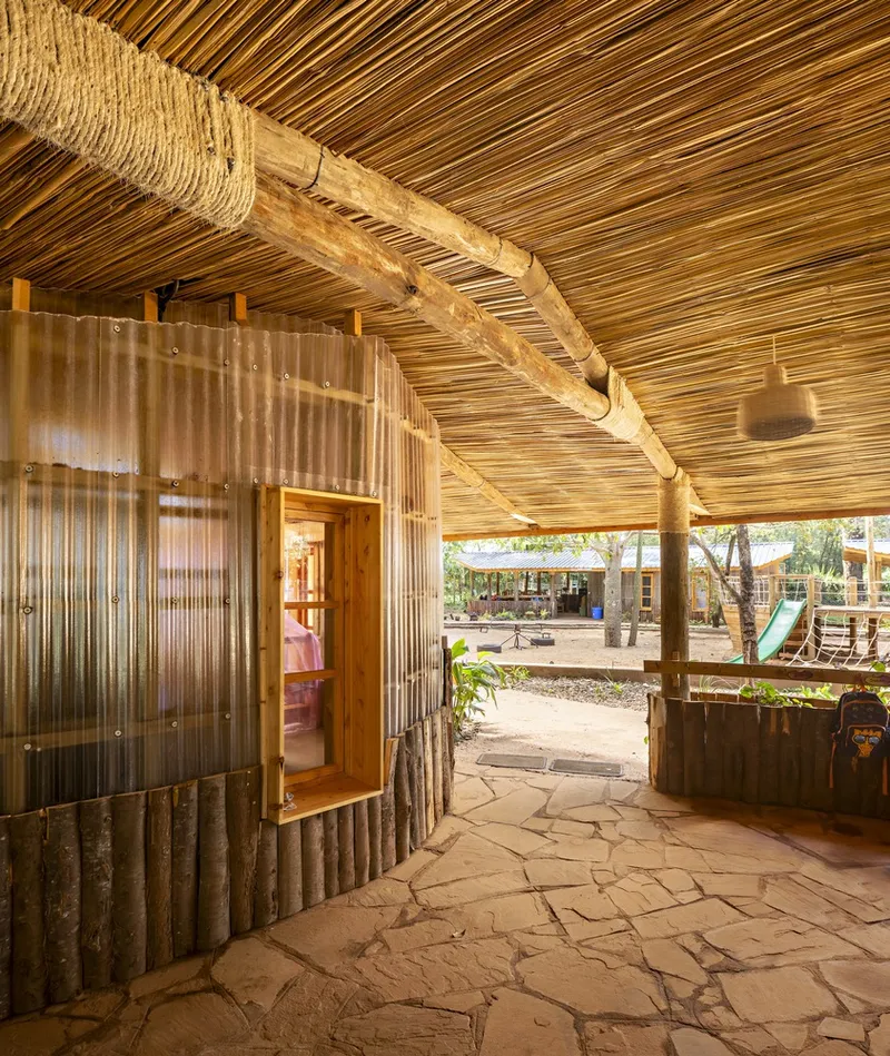 Covered corridor with bamboo slat ceiling, rope-wrapped timber rafters, and corrugated metal wall with open doorway