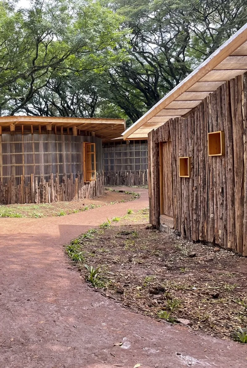 Curved pathway between timber pavilions clad in vertical logs with small window openings beneath overhanging trees