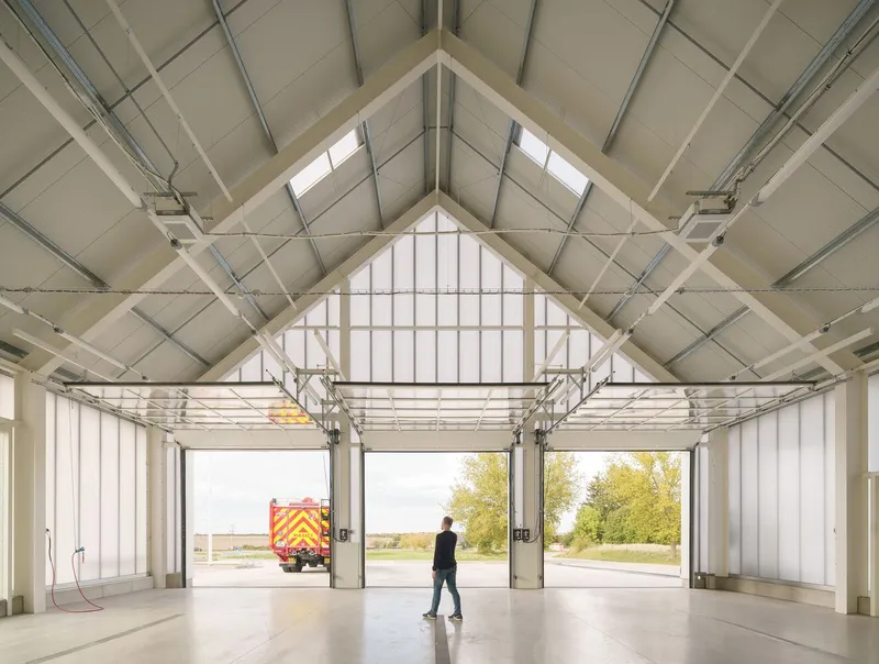 Interior of a fire station garage bay with exposed gabled roof structure and person walking toward open doors