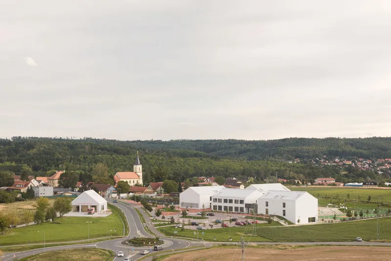 Aerial view of three white gabled volumes adjacent to a village with church spire and forested hills