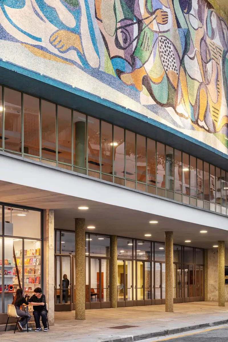 Exterior covered walkway with circular columns beneath a mosaic mural frieze and glazed upper level