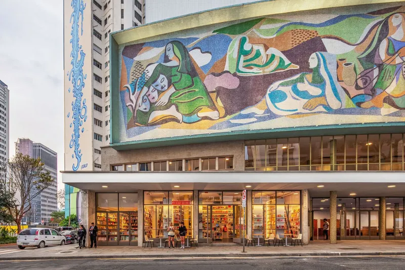 Street-level view of the facade with mosaic mural and pedestrians seated at outdoor tables at dusk