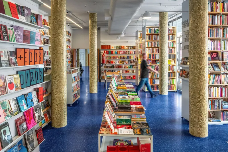 Bookstore interior with cork-clad columns, blue terrazzo flooring, and a figure browsing shelves