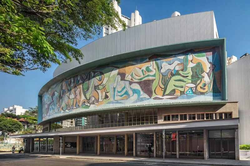 Curved facade with mosaic mural above a colonnade of slender columns framed by leafy trees