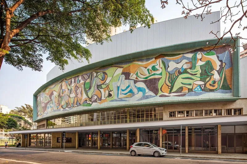 Street facade with curved canopy bearing colorful figural mosaic beneath overhanging trees at golden hour