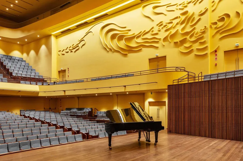 Concert hall interior with relief-sculpted wall, tiered seating, vertical wood slat panels, and grand piano on wooden stage