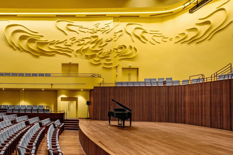 Stage view showing the relief-patterned yellow wall, curved balcony, wood cladding, and grand piano on timber flooring