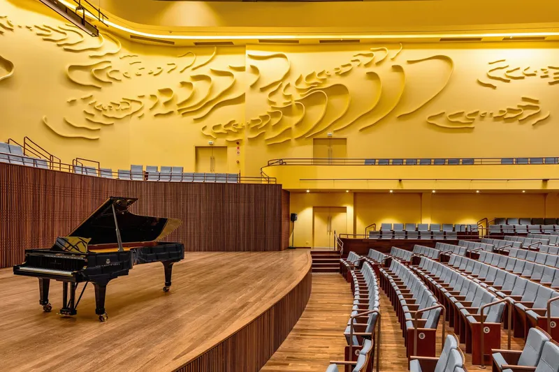 Grand piano on curved timber stage beneath the sculpted yellow wall and auditorium seating under warm lighting