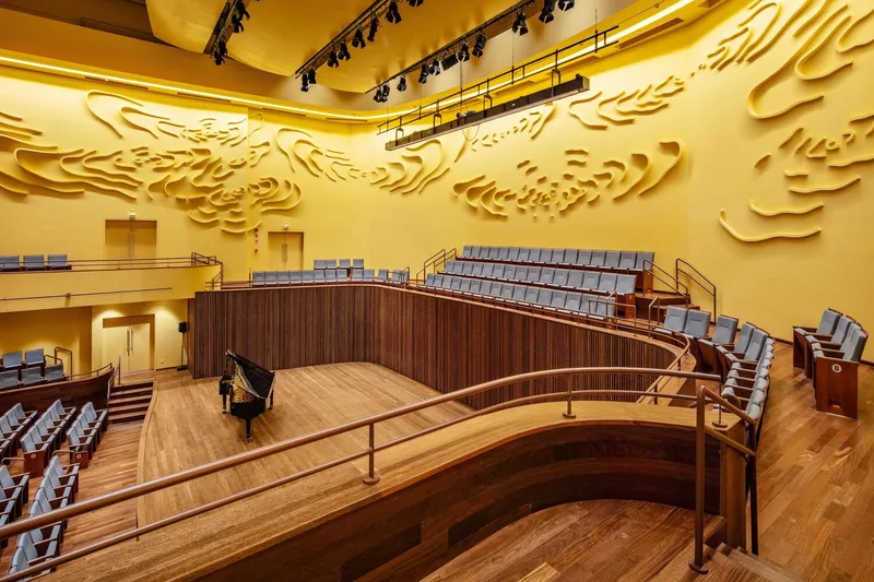 Curving timber stage apron with grand piano beside vertically slatted balcony fronts and relief wall