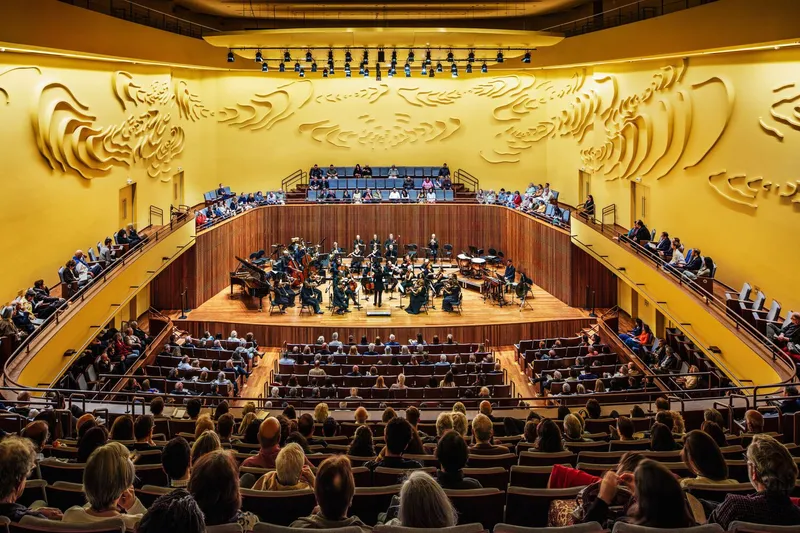 View from rear seating toward orchestra on stage framed by curved timber balcony and relief wall