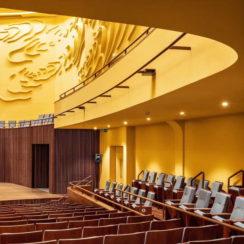 Curved balcony edge below recessed ceiling with relief panels and timber screen beside seating rows