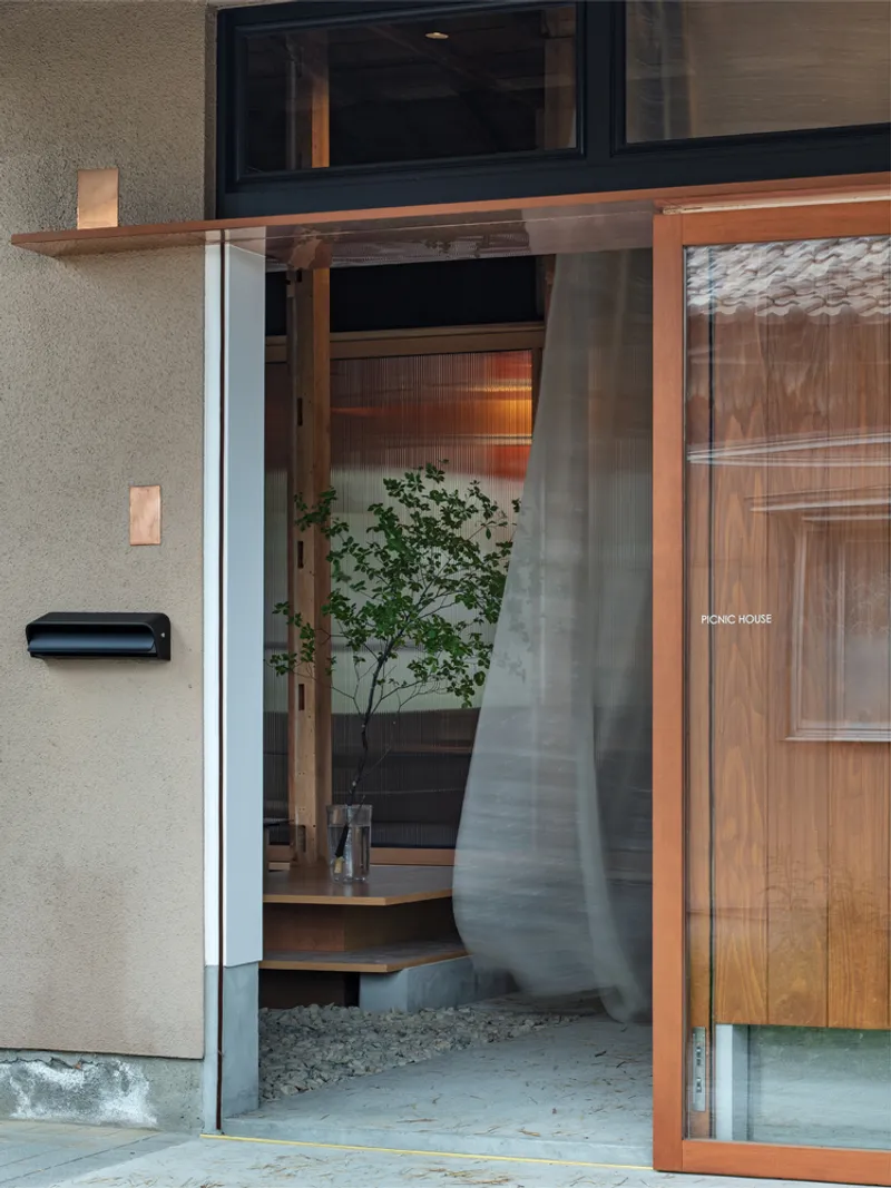 Glass entrance door framing view of interior timber platform with potted tree and gravel floor