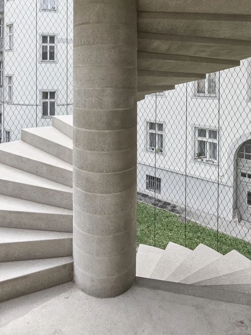 Spiral concrete stair wrapping a cylindrical column overlooking a mesh-screened courtyard with grass