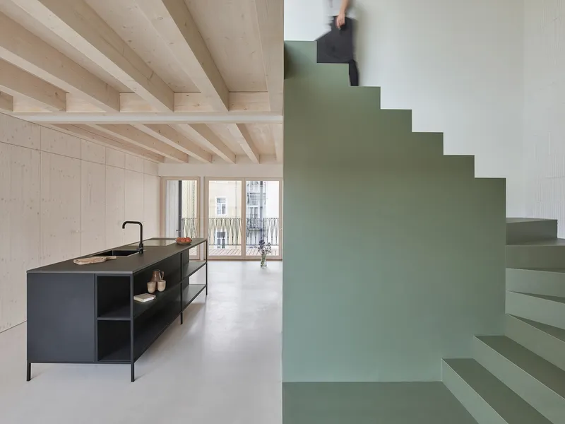 Open kitchen with black island beneath exposed timber joists and a person ascending the sage stair