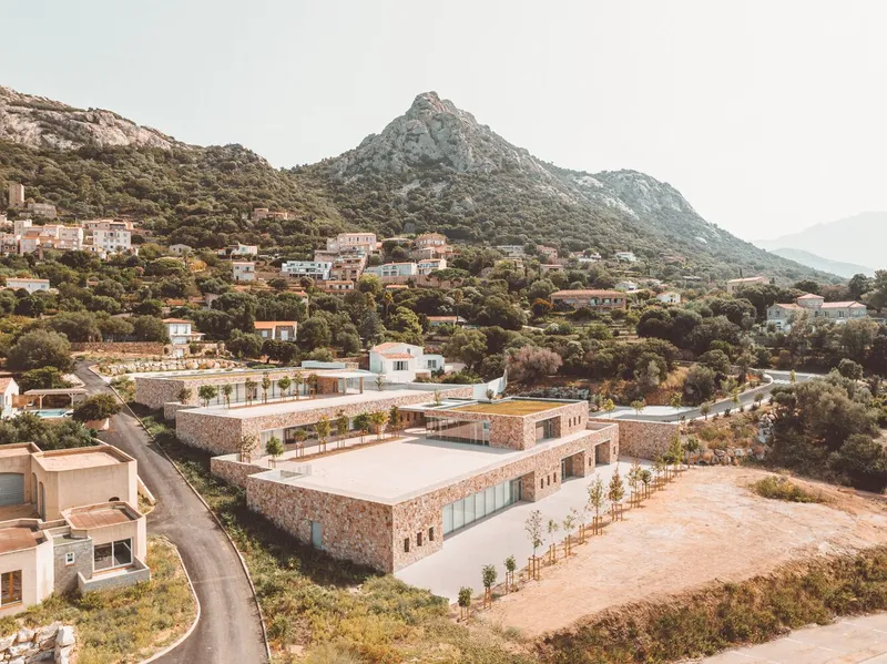 Aerial view of the low-lying stone-clad volumes nestled into the hillside beneath a rocky peak