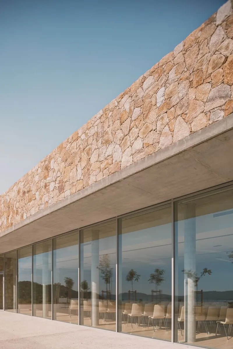 Stone parapet wall above full-height glazing enclosing dining space with chairs and trees