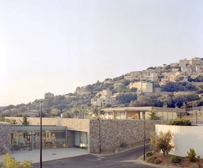 Stone-clad volumes with flat roofs nestled below a hillside village in afternoon light