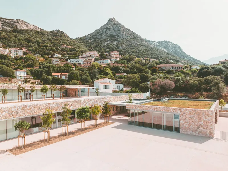 Courtyard with pink stone walls, glass corners and green roof beneath hillside village and mountain