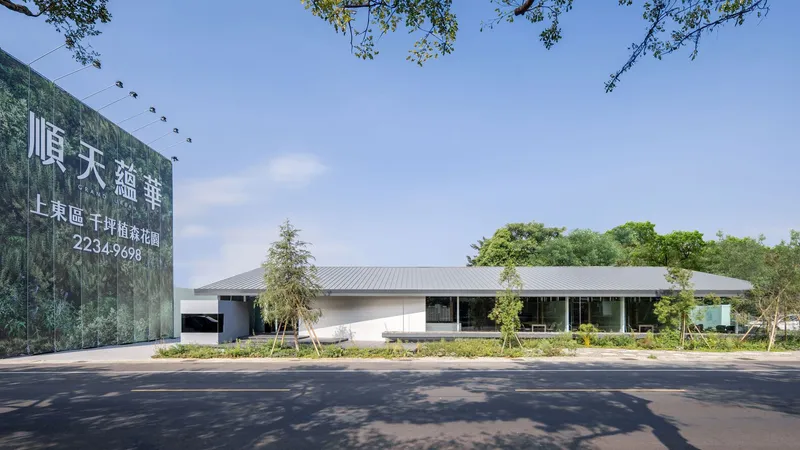 Street view of the low-slung pavilion with corrugated metal roof and landscape beds under dappled tree shade