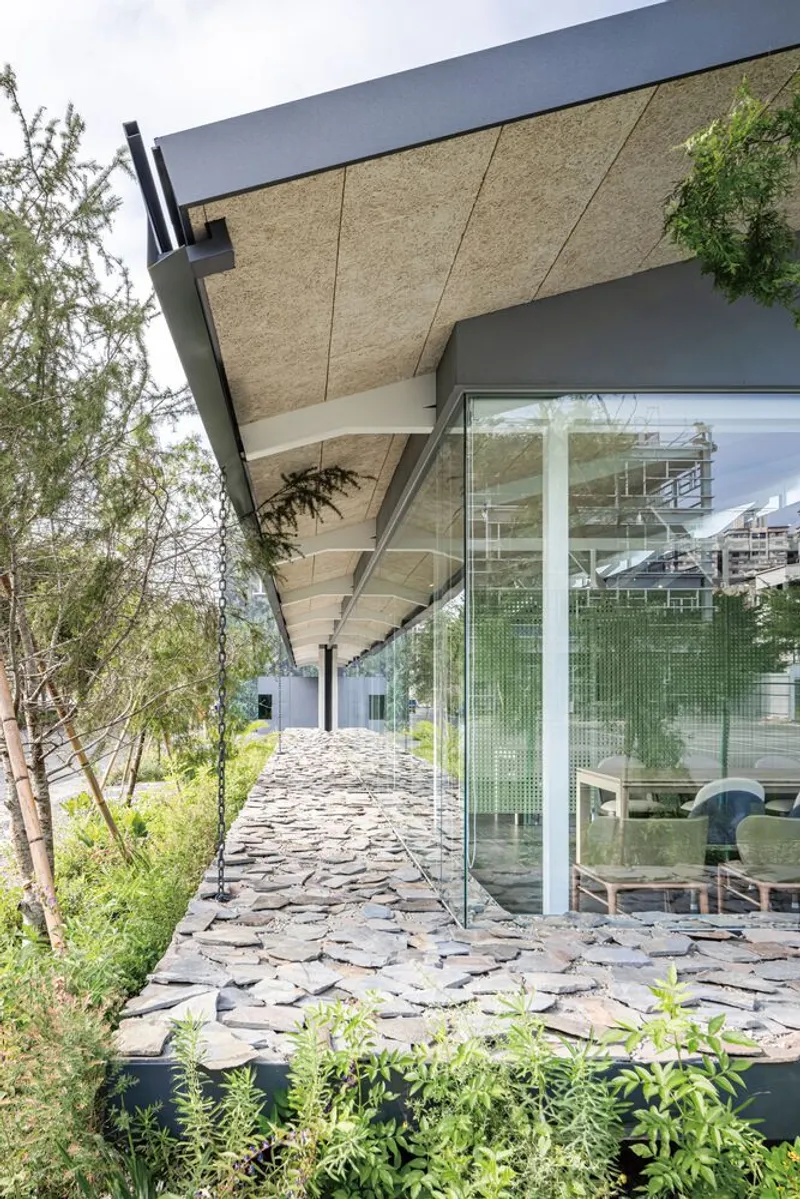 Covered walkway with stone pavers alongside glass facade reflecting planted trees and exposed ceiling structure