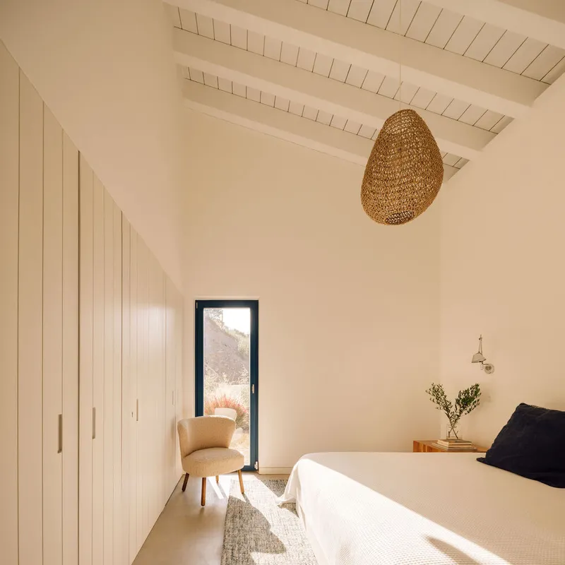 Bedroom with exposed timber ceiling beams, vertical panel wardrobe and narrow window framing a distant hillside