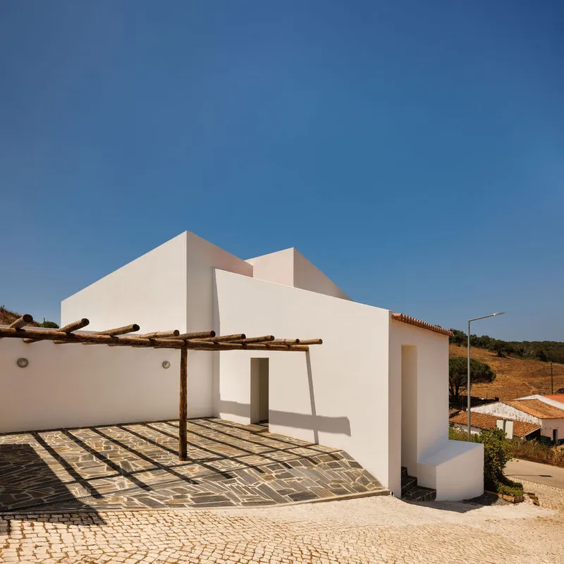 White stucco facade with timber pergola casting shadows over cobbled paving under clear blue sky