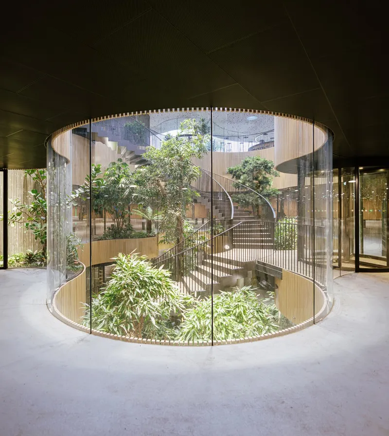 Cylindrical glass atrium enclosing planted courtyard and curved staircase with timber screens