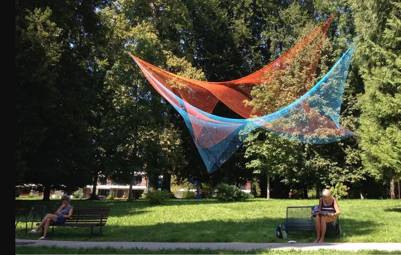 Overhead canopy of orange and blue mesh netting suspended above park benches on a sunny afternoon