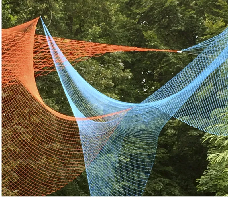 Suspended tensile mesh canopy in orange and blue gradients stretched between trees in dappled daylight