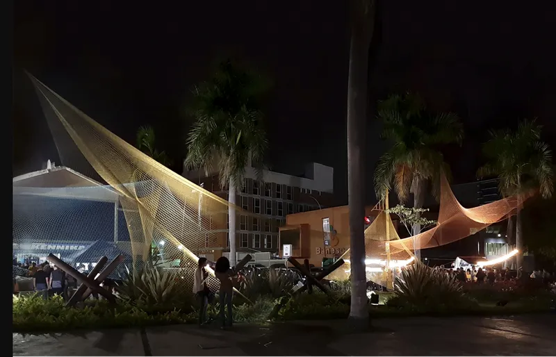 Tensile mesh installation lit at night over a public plaza with palm trees and pedestrians