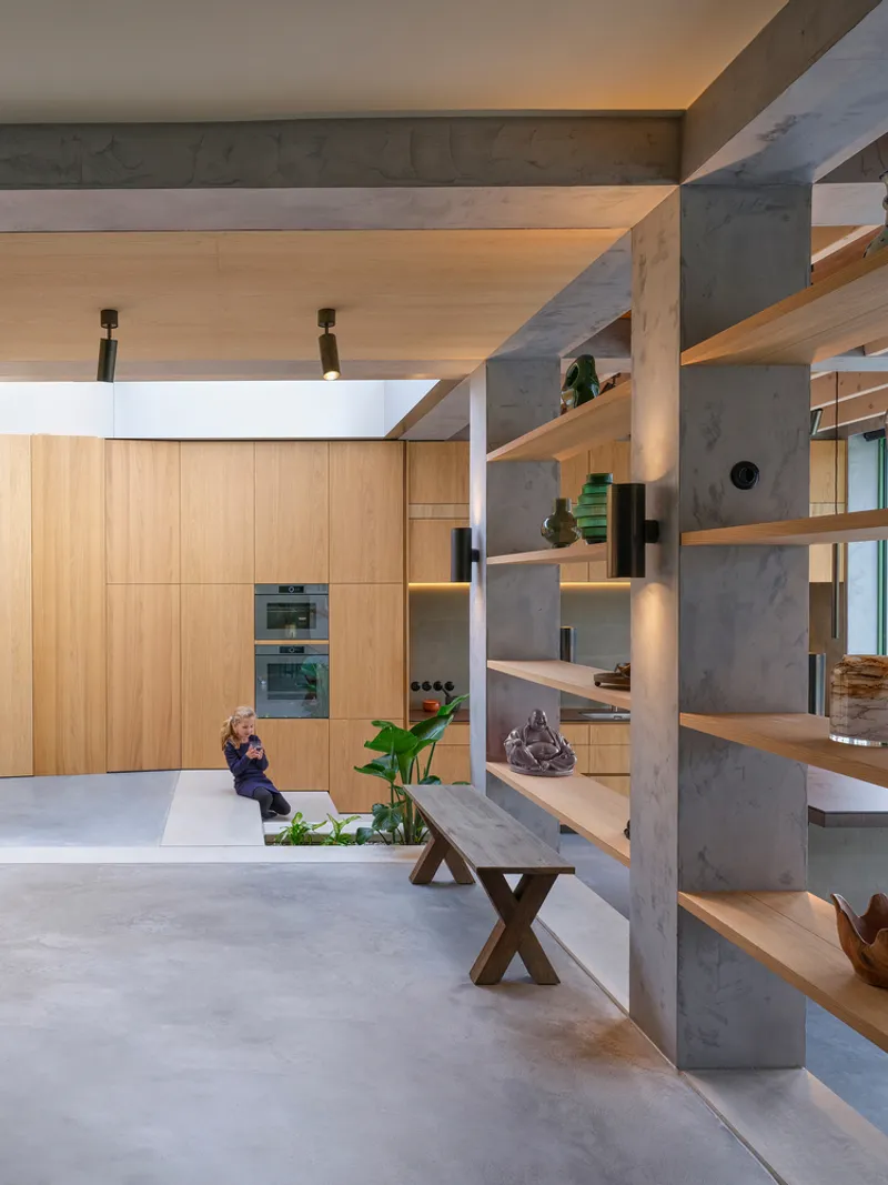 Open-plan interior with concrete shelving columns and oak cabinetry illuminated by warm ceiling light