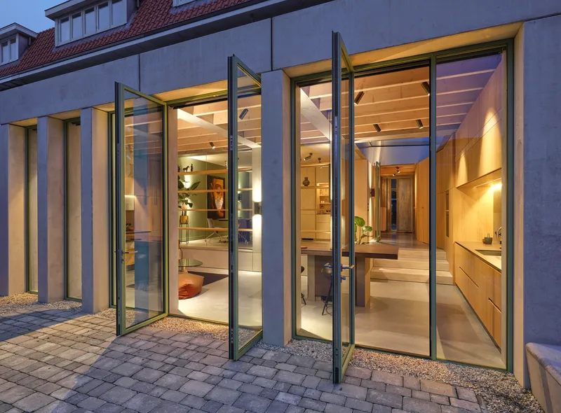 Folding glass doors opening from a paved courtyard into the illuminated interior at dusk