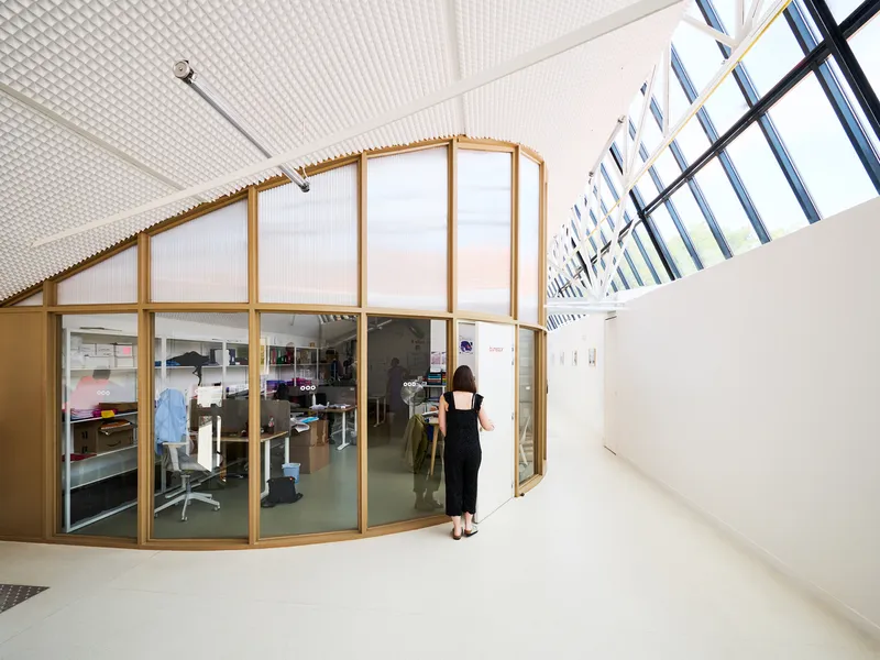 Curved glass and timber partition enclosing workspace beneath a gabled skylight with white truss structure