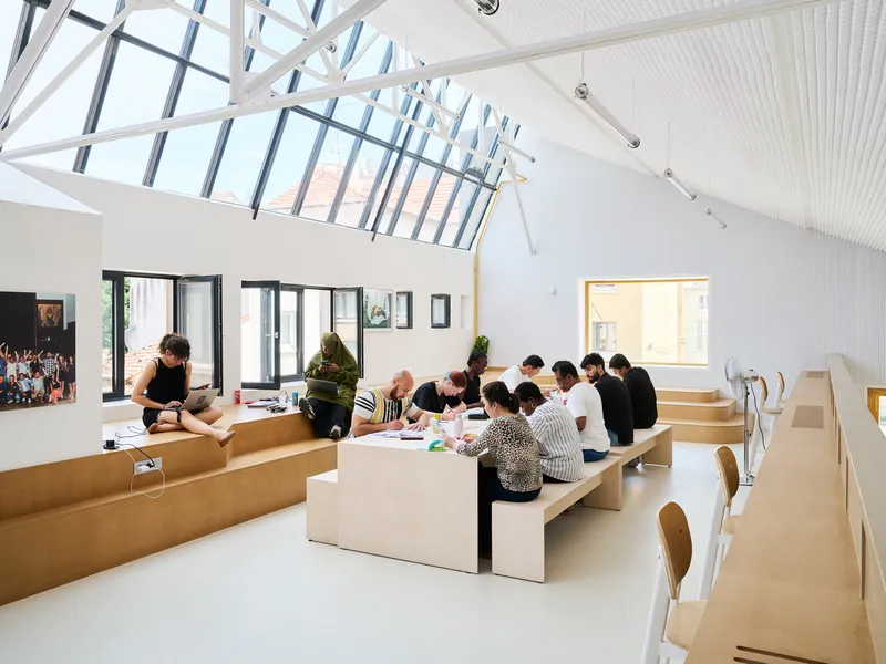 Vaulted interior with exposed steel truss skylight where participants gather at tiered plywood seating platforms