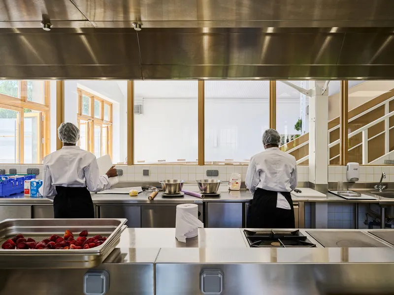 View from the stainless steel prep counter toward timber-framed windows overlooking the adjacent dining space