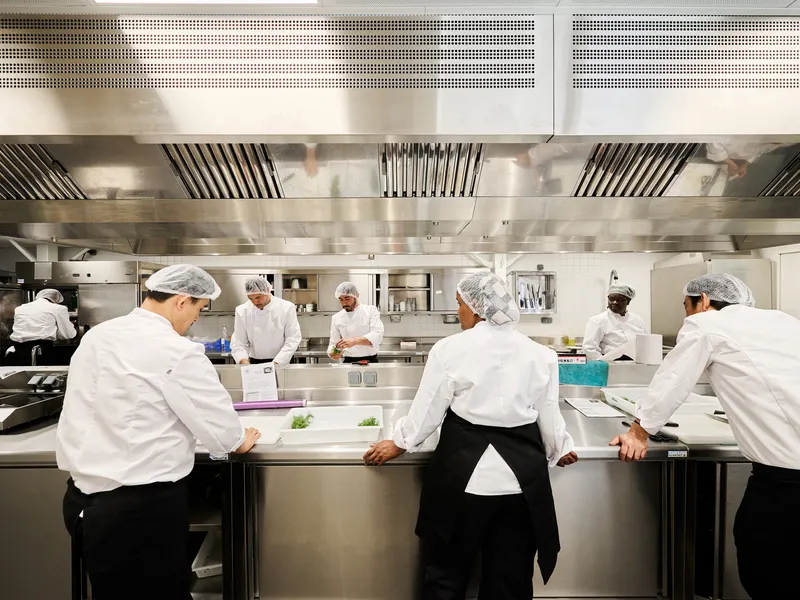 Commercial kitchen with stainless steel counters and overhead exhaust hoods while staff in whites prepare food