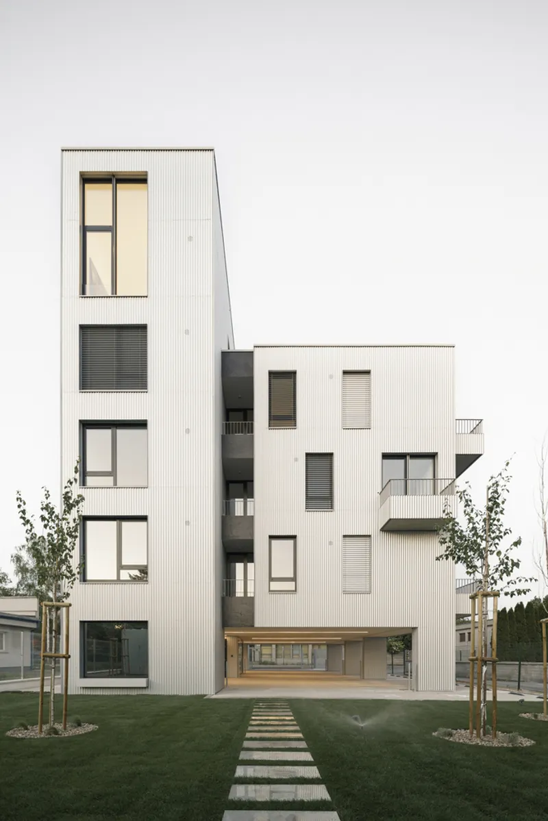 White corrugated metal facade with varied window openings flanked by young trees and concrete stepping stones