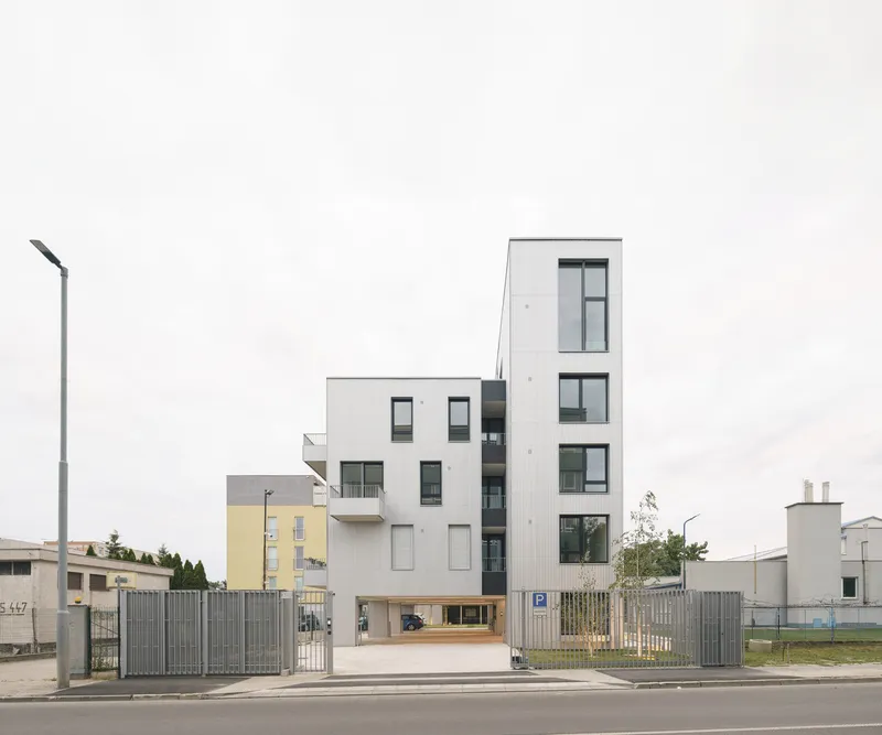 Street view of a white tower with recessed balconies rising from a timber-clad ground floor under an overcast sky