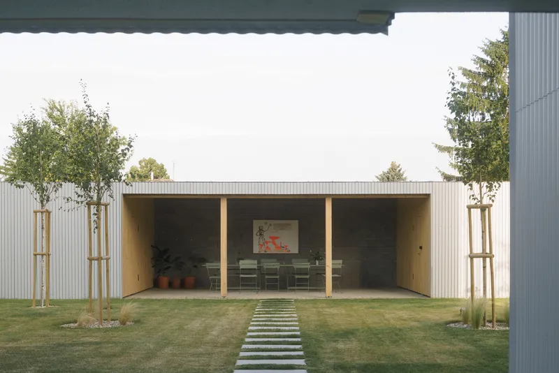 Outdoor pavilion with timber posts and corrugated metal cladding framed by young trees on a lawn